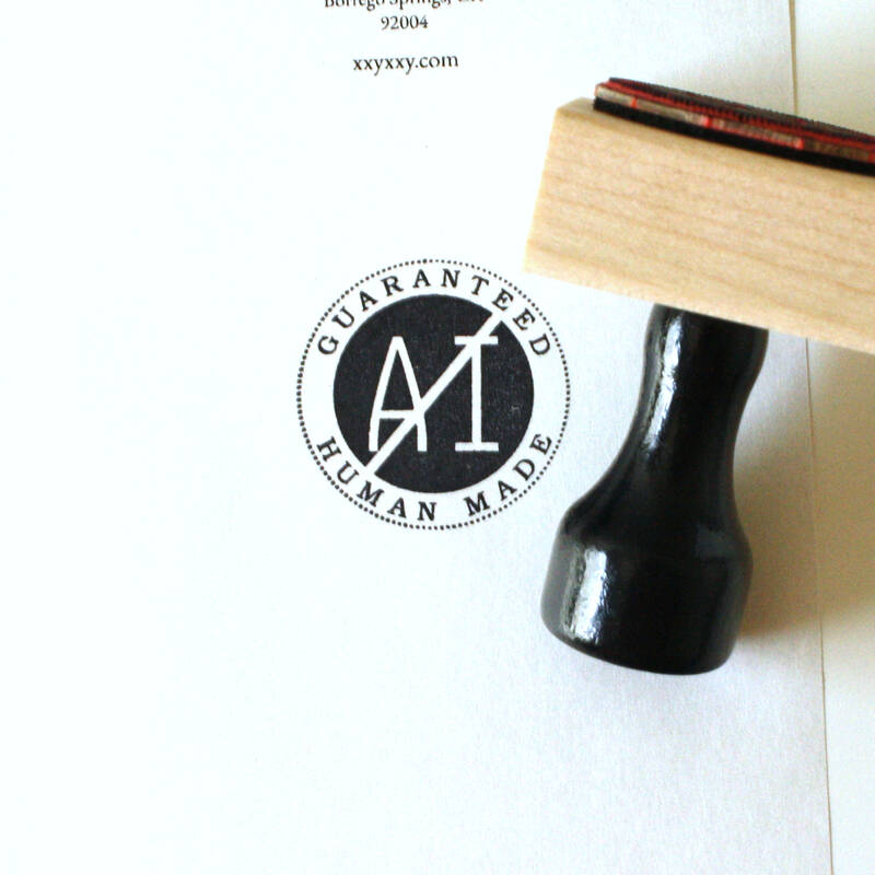 A wood base, rubber stamp, with black handle, laying on its side next to an imprint which reads Guaranteed Human Made in a circle surrounding a black circle with AI in the center crossed out.